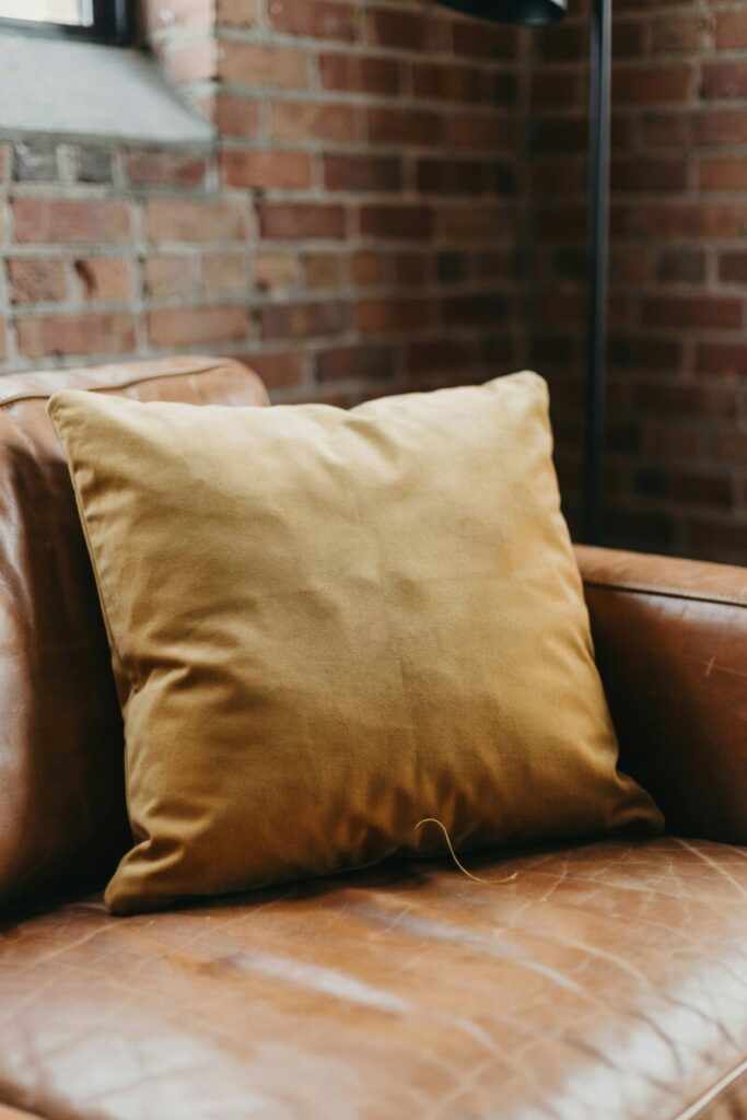 Mustard yellow velvet throw pillow on a cognac leather sofa with exposed brick wall in the background