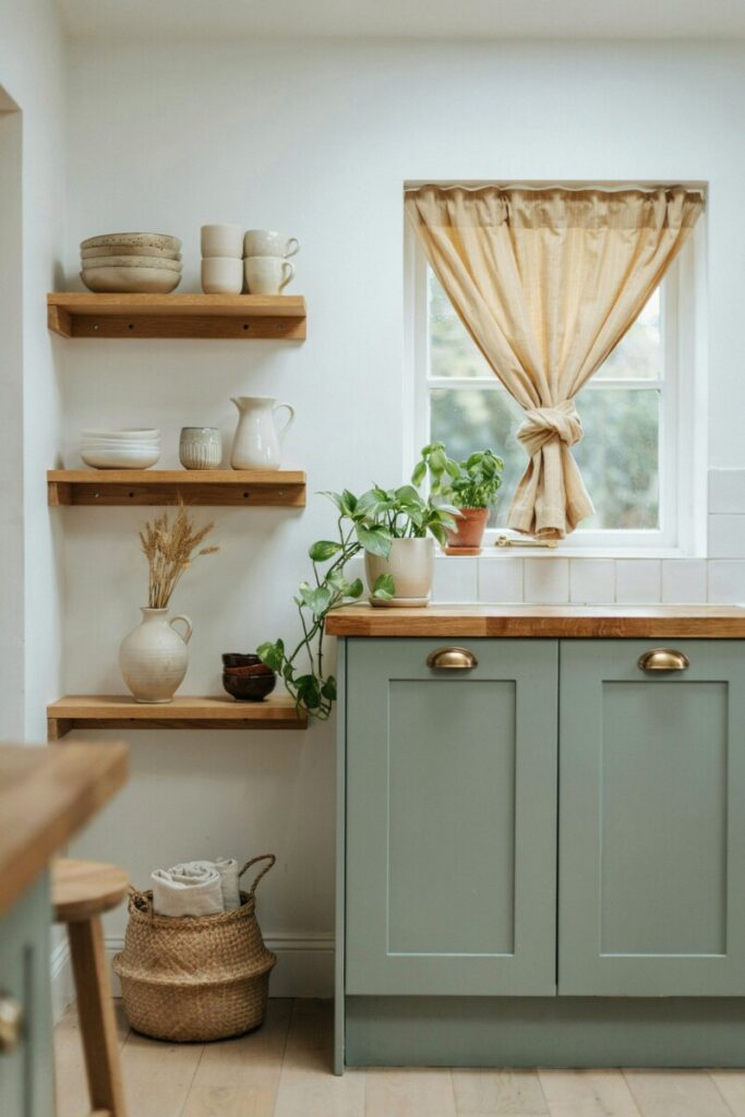 Sage green kitchen cabinets with open wood shelving and cream ceramics showcasing a soft meadow green cottagecore color palette.